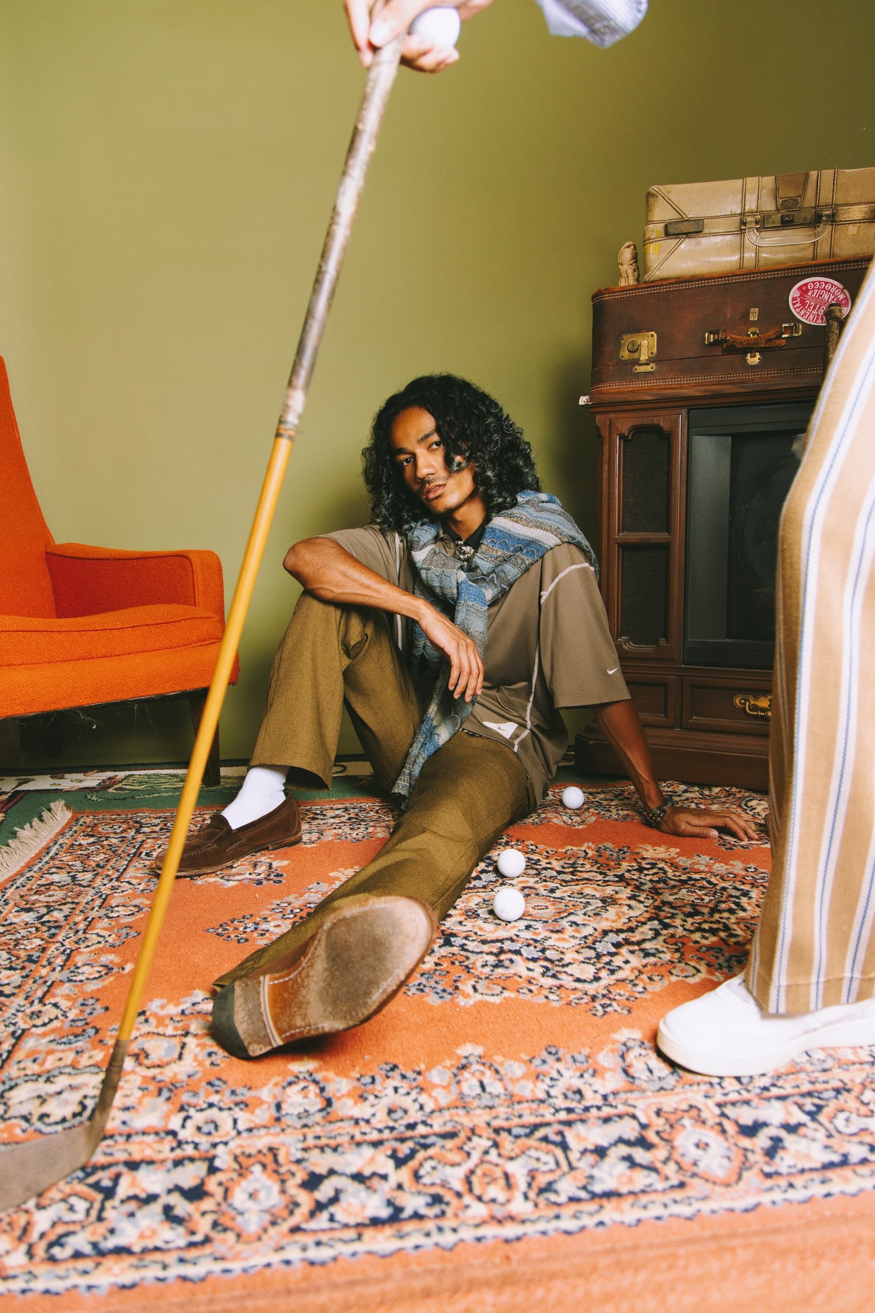 A model seated on a vintage rug holding a wood-shafted golf club, wearing pieces from the Staples Collection