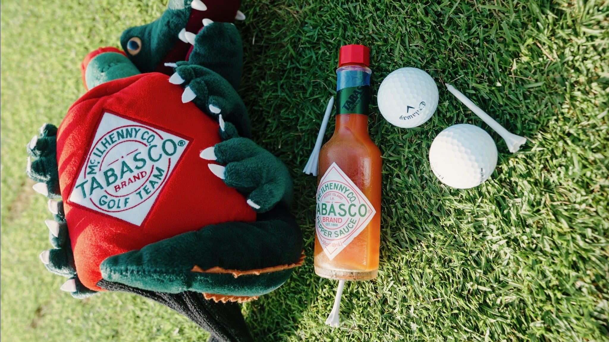 A vintage Tabasco Golf Team gator headcover next to a bottle of Tabasco pepper sauce, golf balls, and tees on cut grass