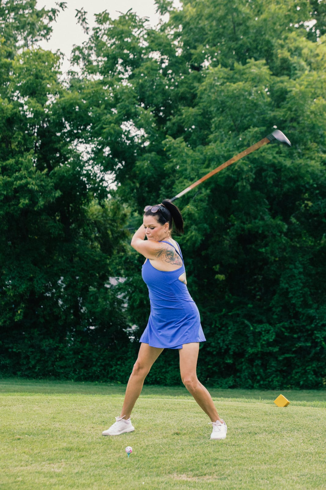 Golfer in a blue dress at full extension on her follow-through, dark trees behind her