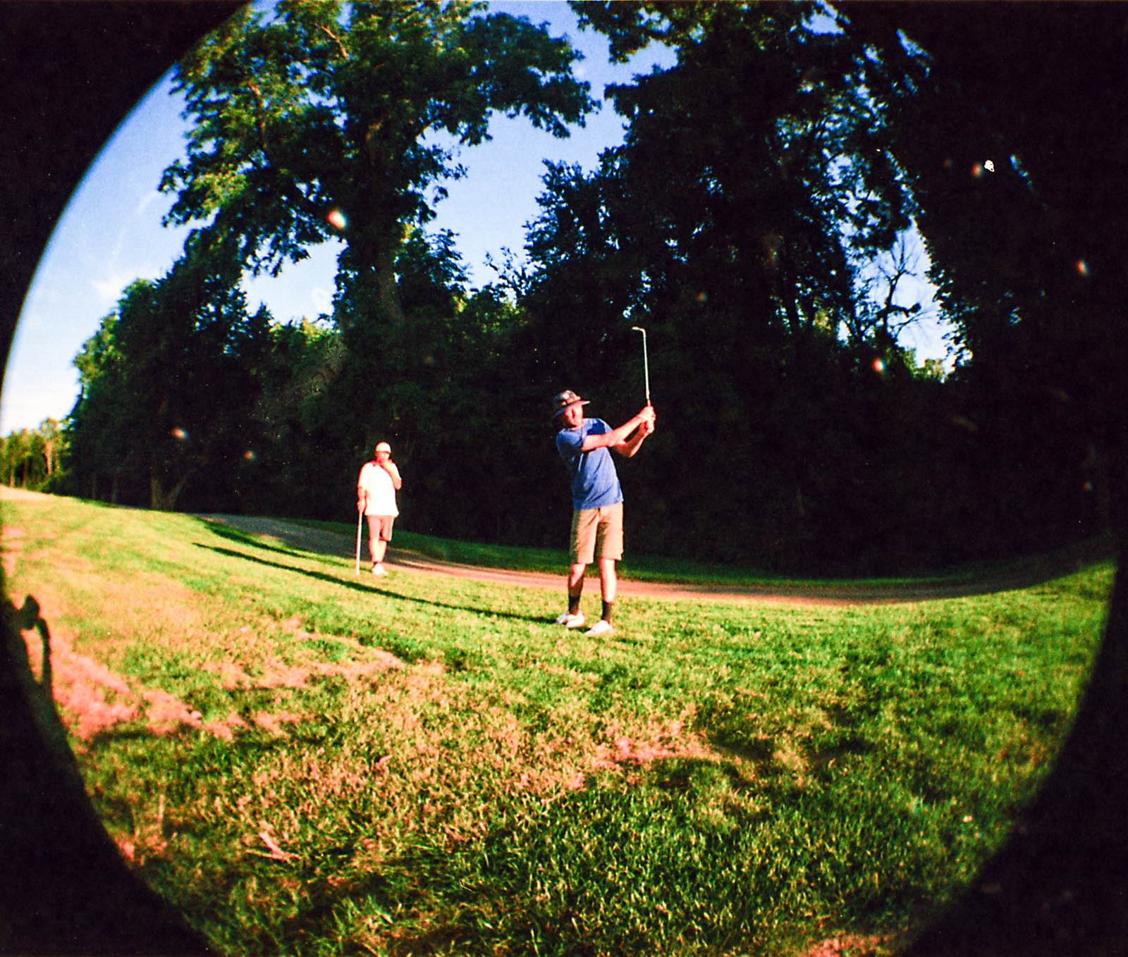 Golfer's follow-through in the shade of the tree line, a partner watching down the fairway