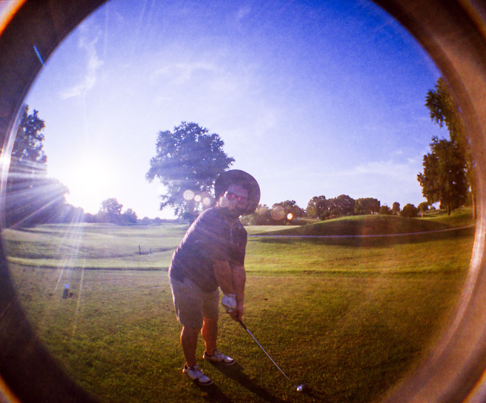 Golfer in a wide-brim hat addressing the ball as the sun flares low over the course