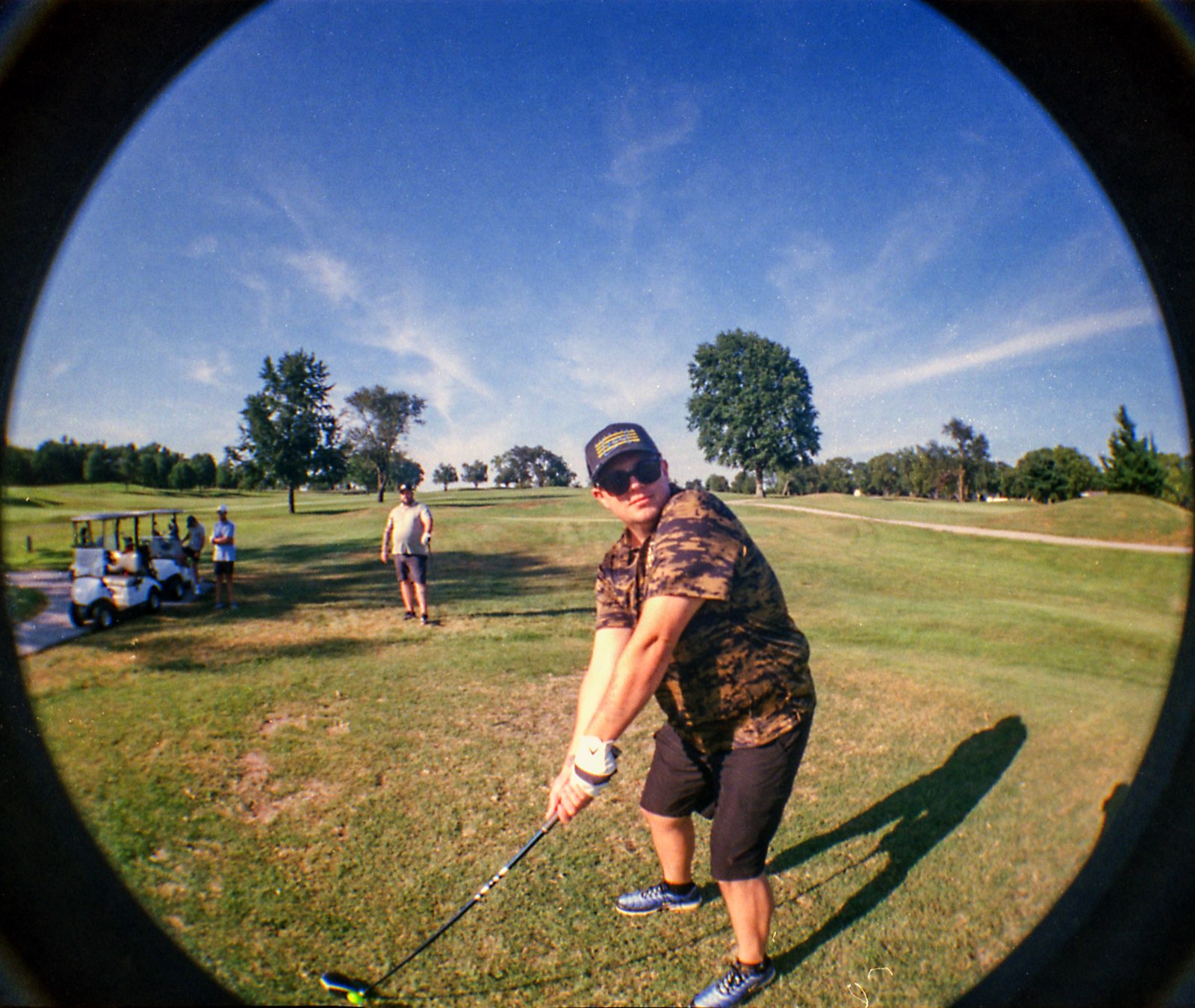 Golfer in a camo tee and shades following through, the group gathered by the carts behind him