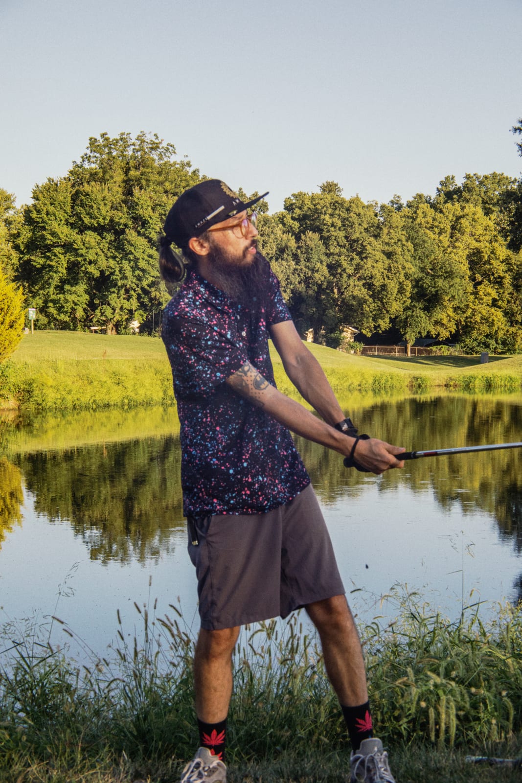 Bearded league member in a patterned polo posing with his club beside the pond
