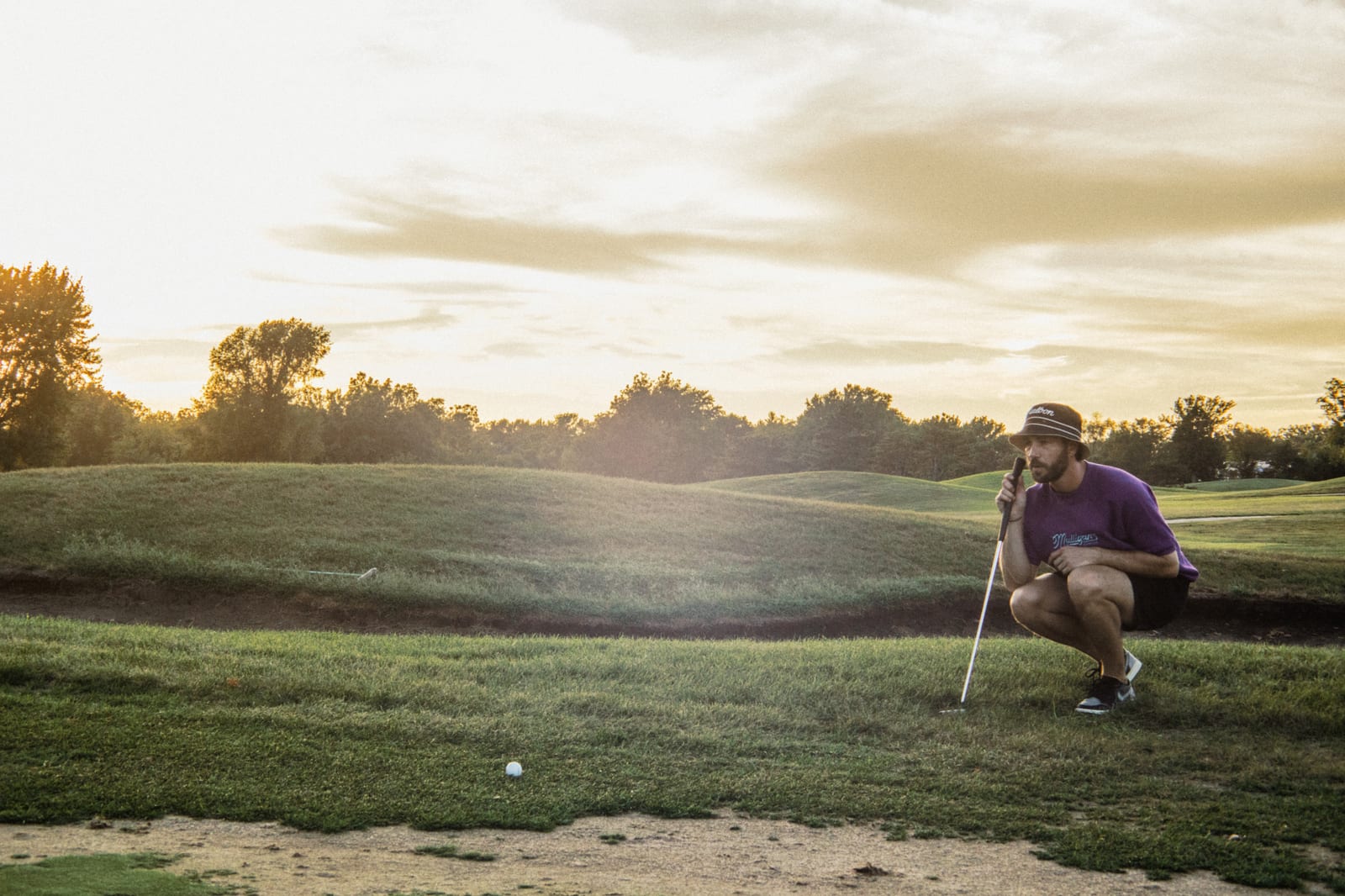 Golfer in a purple tee crouched reading a putt under a streaked sunset sky