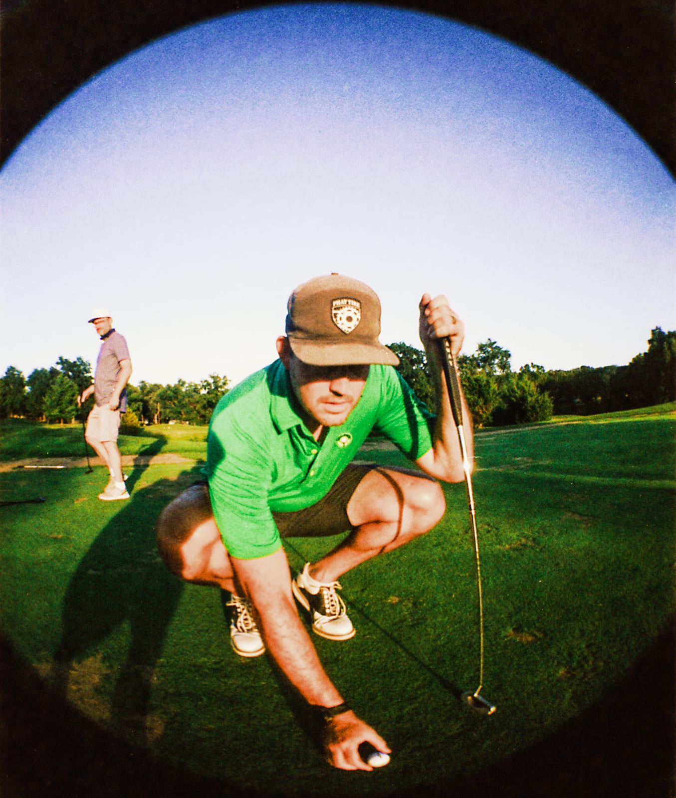 Golfer in a green polo crouched behind his putter, grinning into the fisheye